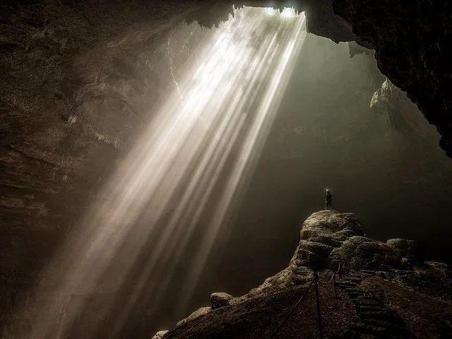 Person standing on a rock inside a cave illuminated by a strong light beam.