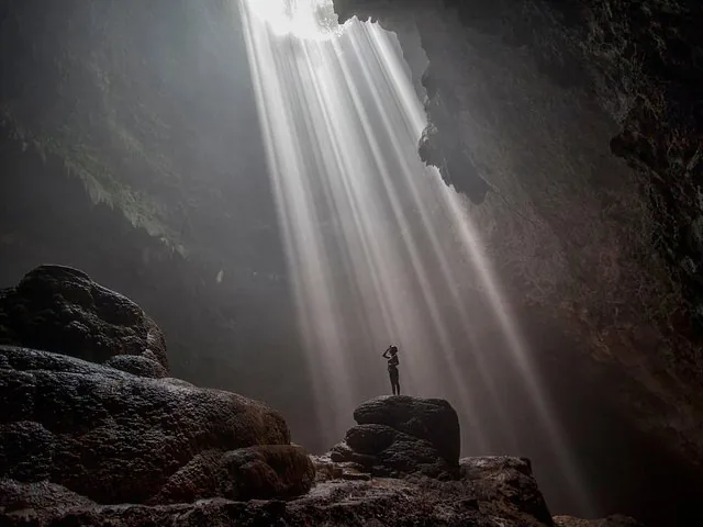 Person standing on rocks in a cave looking up at intense light beams.