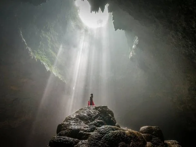 Person in red dress standing on a rock under a light beam in a cave.