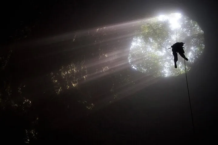 Person rappelling down into a cave through a large opening illuminated by sunlight.