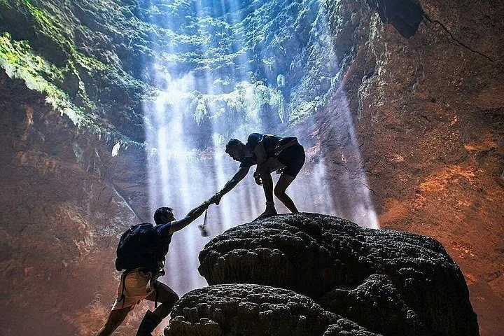 Two people climbing rocks in a cave under a beam of sunlight.