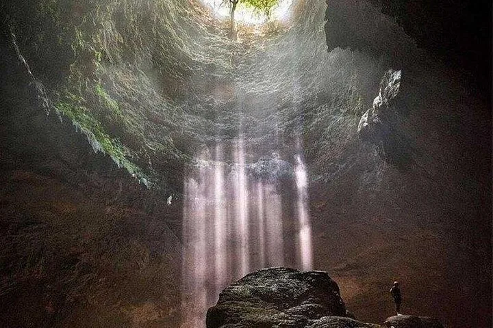 Person standing on a rock in a cave looking up at a light beam.