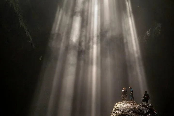 Three people standing on a rock inside a cave under bright light beams.