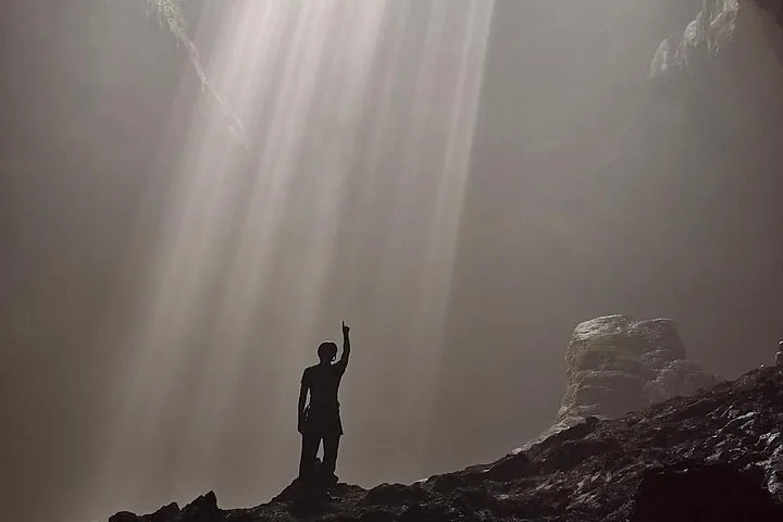Silhouette of a person pointing up towards a light beam inside a cave.